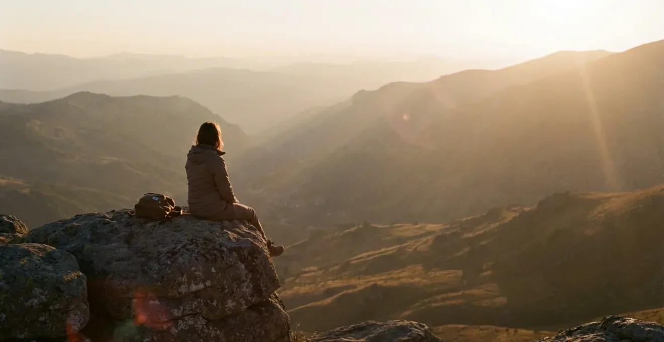 Femme contemplative assise seule sur un rocher face à un panorama montagneux au coucher du soleil, symbolisant la quête d'inspiration et le ressourcement